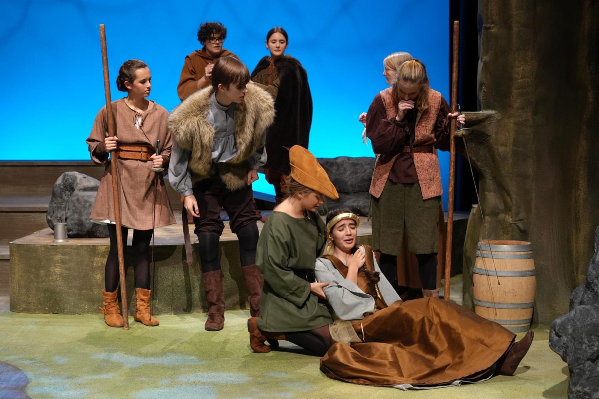 Marian/Robin Hood, played by Lainie Beamer (‘25), comforts Lady Shirley, played by Leela Zaveri-Tabb (‘25), at the Merry Men’s homebase in the Sherwood Forest. Behind them are more Merry Men: Alanna Dale (Zara Zierhut (‘27)), Little John (Jackson Weisser (‘26), Will Scarlett (Bex Balsdon (‘26)), Friar Truck (Dash Flaig (‘27)), Much the Miller’s Son (Madison Gasaway (‘28)), and Tommy of No Consequence (Sydney Whitman (‘26)).  